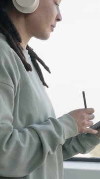Vertical video: Panning adult drawing on pad with stylus at window in sage top, headset, copy space