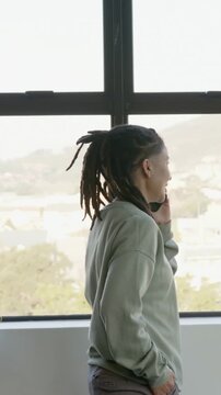 Vertical video: Shifting, nonbinary person lifting phone and calling at home window plant green top