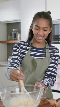 Vertical video: Smiling teen in green apron whisking batter in bowl in home kitchen, adult guiding