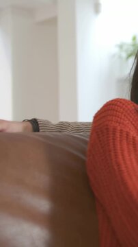 Vertical video: Settling woman in orange knit shifting on couch for comfort with watch, copy space