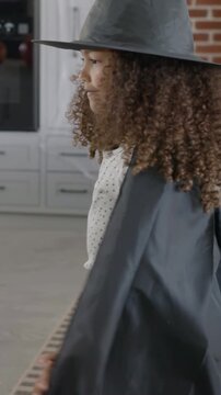 Vertical video: Smiling mom in sweater reaching placing witch hat cape on kid at home holding pail
