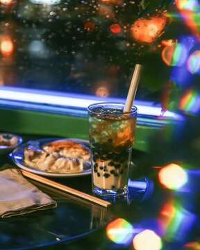 Iced milk tea with boba pearls and steamed dumplings on a dark table, glowing under neon lights by a rainy window for cafe and food branding