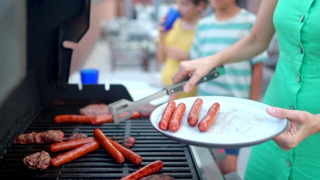 Mother grilling hot dogs and burgers on a barbecue for a summer family cookout, using tongs to place cooked sausages on a plate while children wait in the background