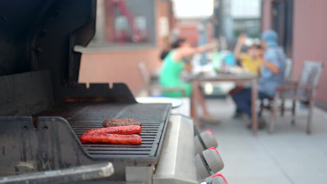Summer barbecue with hot dogs and hamburgers cooking on an outdoor grill, while a group of friends enjoy a backyard party in the blurred background on a sunny day