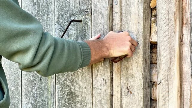Close up of a hand pulling shut the weathered wooden door of a rustic woodshed filled with stacked logs. Country life security and firewood storage concept.
