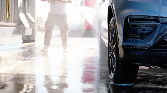 Worker cleans car with high-pressure water jet. Sunlight reflects off wet pavement and glossy vehicle. Water sprays forcefully against dark tires and bumper. Person moves deliberately