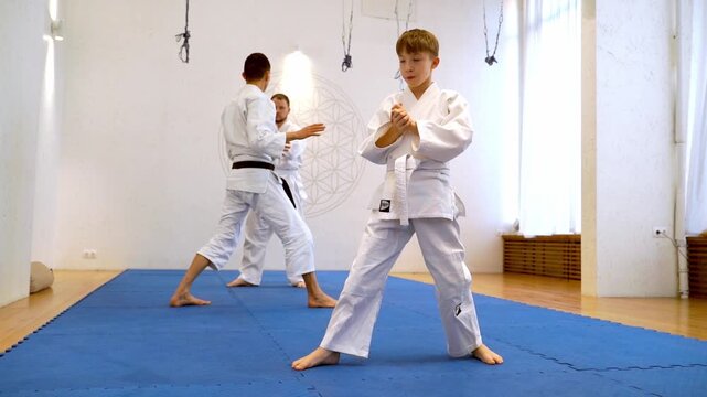 Two martial artists practice judo in a bright dojo. One performer executes a move while the other watches attentively. Young practitioners train in coordinated movements on a blue mat