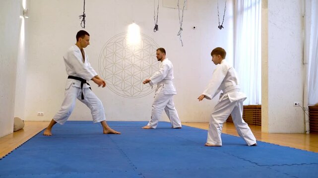 Three martial artists practice in a bright dojo. One instructor guides two students with calm focus. The blue mat contrasts with clean white walls. Participants move with discipline and respect