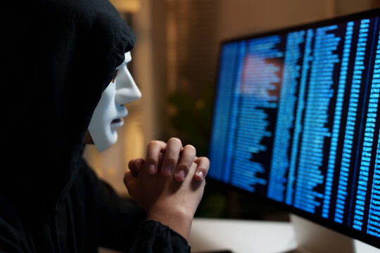 Anonymous hacker wearing a white mask and typing code on a computer, representing cybercrime, data breaches, and digital security threats.