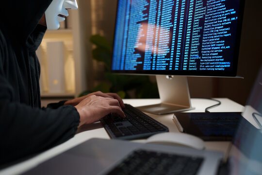Anonymous hacker wearing a white mask and typing code on a computer, representing cybercrime, data breaches, and digital security threats.