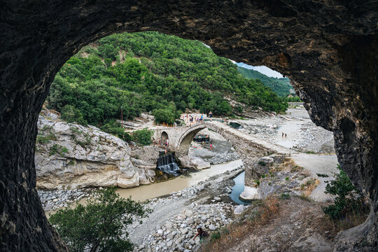 Ancient stone arch bridge viewed through a cave opening in a green canyon landscape near Benja thermal baths, Albania.
