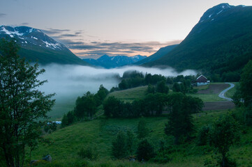 Low sunset fog drifts over a green valley with farms and forests beneath steep mountains. © Sergio