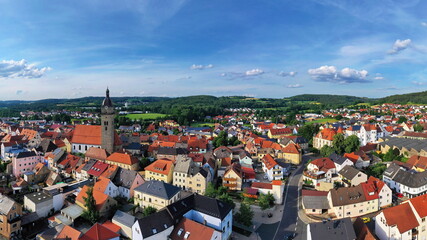 Obraz premium Malerische Luftaufnahme von Auerbach in der Oberpfalz mit der markanten Stadtpfarrkirche, traditionellen roten Dächern und idyllisch grüner Landschaft im Hintergrund unter blauem Himmel.