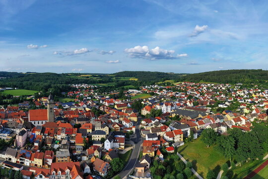 Malerische Luftaufnahme von Auerbach in der Oberpfalz mit der markanten Stadtpfarrkirche, traditionellen roten D&auml;chern und idyllisch gr&uuml;ner Landschaft im Hintergrund unter blauem Himmel.