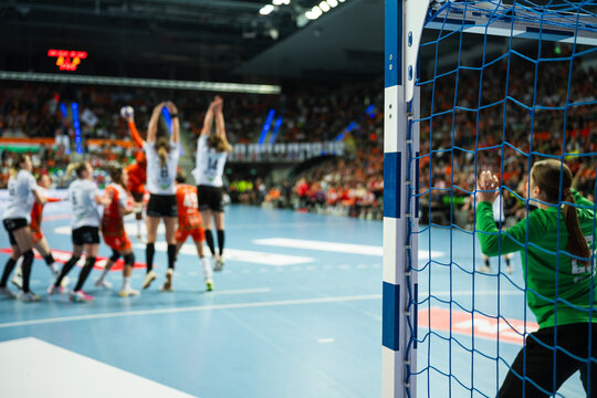 Handball goalkeeper defending the goal during a professional indoor match, viewed through the goal net with intense game atmosphere