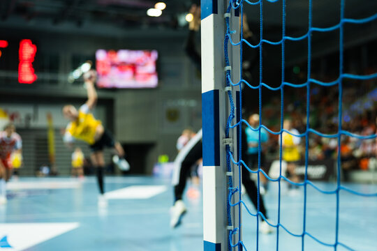 A blurred view of a live handball match seen from behind the goal post and blue net, with players and referees in action.