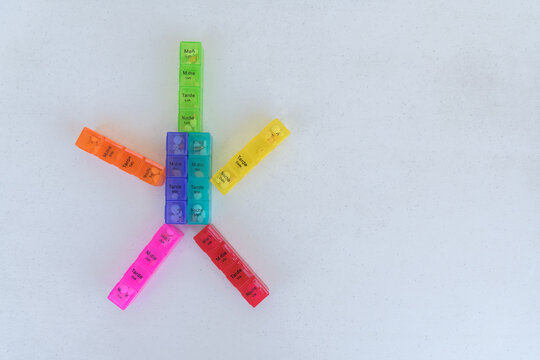 Seven colorful pill organizers form a pinwheel or star-shaped pattern, with a central 2x2 block core and arms radiating outward, leaving open space to the right.