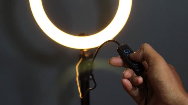 Hand of an Asian man adjusting the brightness of a glowing LED ring light using an inline controller, with illuminated control buttons against a dark background.