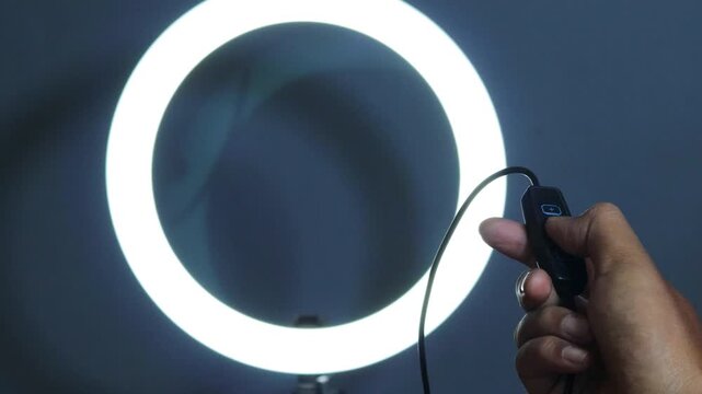 Hand of an Asian man adjusting the brightness of a glowing LED ring light using an inline controller, with illuminated control buttons against a dark background.