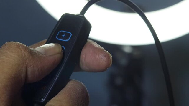 Hand of an Asian man adjusting the brightness of a glowing LED ring light using an inline controller, with illuminated control buttons against a dark background.
