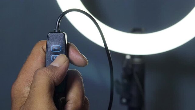 Hand of an Asian man adjusting the brightness of a glowing LED ring light using an inline controller, with illuminated control buttons against a dark background.