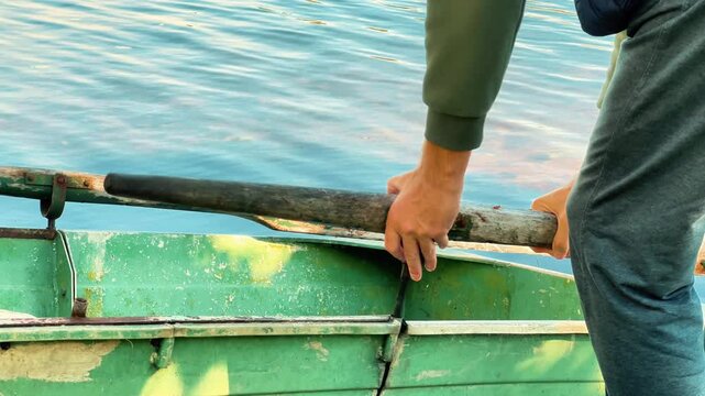 Close up view of a man installing a wooden oar into the metal oarlock of a green rowing boat on a calm lake. Preparing for a boat trip and leisure activity.