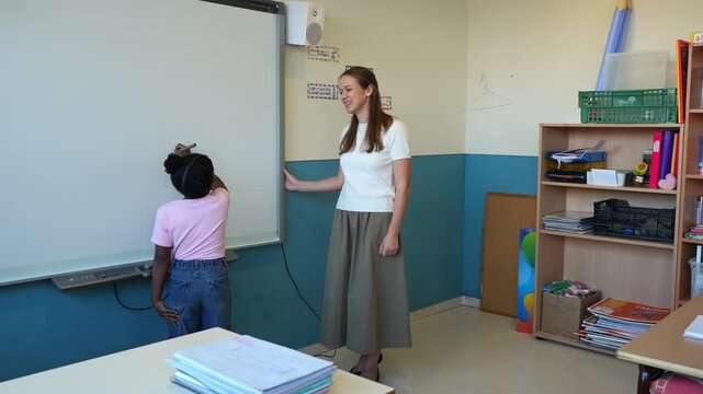 Young female teacher giving instructions to a little african american student writing on the whiteboard. Primary school class with a female mentor