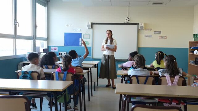 Young female teacher standing in front of a class during a lesson, while a young pupil raises their hand to ask or answer a question