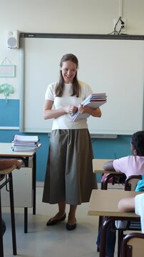 Smiling teacher with notebooks helping a young student at his desk. Happy female educator in a bright elementary school classroom