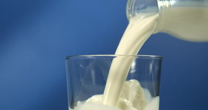 Milk pouring from bottle into glass on blue background in slow motion