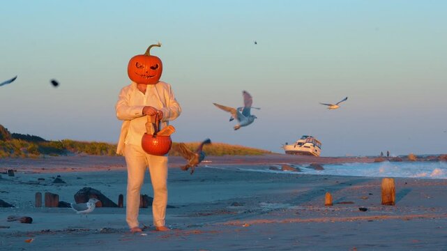 Man wearing pumpkin mask and holding bread basket among gulls. A barefoot figure in a white suit stands on sand with flying birds, waves and a boat behind him.