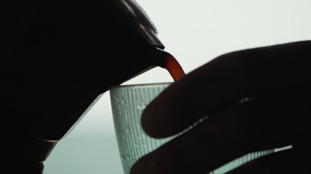Fresh caffeine infusion flows from a brewer into a handheld transparent glass against backlit window light. Extreme close-up shot.