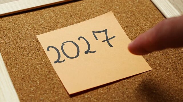 A fingertip slips into the frame toward a peach 2027 sticky note on cork, about to touch or pin it. The moment captures reaching for the coming year, taking action on plans and the first step into 20