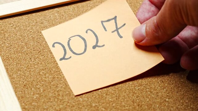 Single peach sticky note with 2027 written in bold black handwriting, stuck slightly askew on a cork pin board between pale wooden frame rails. A simple new year symbol, ready for headers, title card