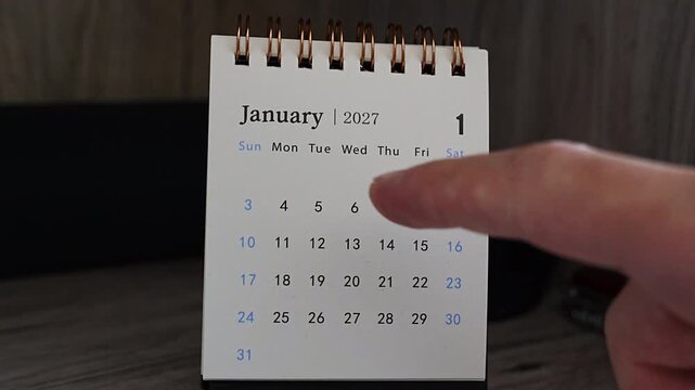 A finger points to January 2027 on a small spiral desk calendar with copper rings, standing on a dark wooden shelf. Ideal for the first month of the year, scheduling appointments, deadlines and new y