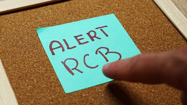 Close up of a cyan sticky note with ALERT RCB handwritten in maroon marker, set at an angle on textured cork. A tight crop for civil defence notices, blackout warnings, crisis readiness and Polish pu