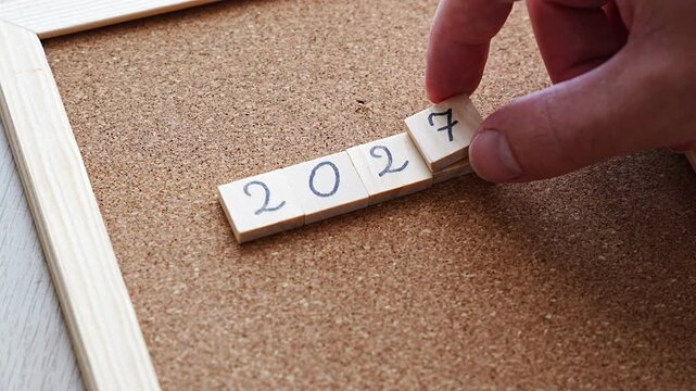 Close view of 2027 laid out in wooden tiles on cork, the new 7 resting on top of the old 6 so a sliver of the previous digit still shows. A layered metaphor for covering the past and stepping into th