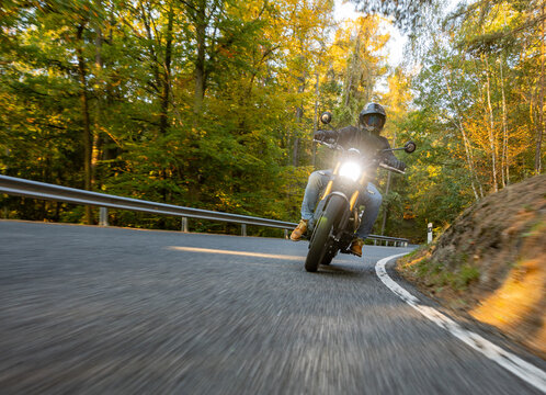 A motorcyclist overtakes a small yellow car on an asphalt road through the forest. The moving shot captures both vehicles in motion against a wooded roadside backdrop.