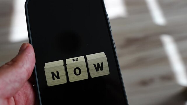 Wooden letter cubes on a smartphone screen flip the middle O so the word NOW becomes NEW, framed tightly with sunlight stripes behind. A sharp concept of innovation, novelty and turning the present m
