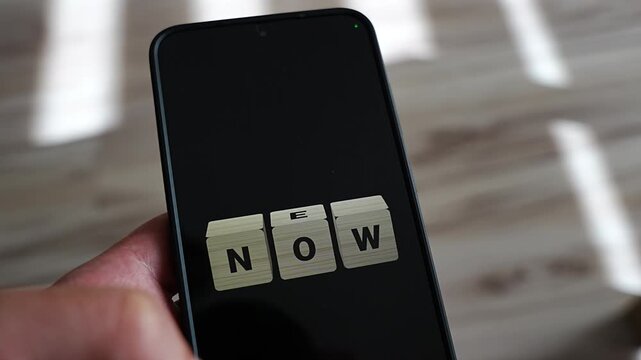 From NOW to NEW, a single wooden cube turns over on an upright smartphone held at the side, with stripes of sunlight on a wooden floor. A symbol of a change of mindset, personal growth, self improvem