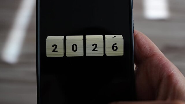 Extreme close up of wooden cubes reading 2026 on a smartphone screen as the last cube rotates to reveal 7, held at the edge by one hand. Macro detail of the year turning, ideal for New Year Eve and 2