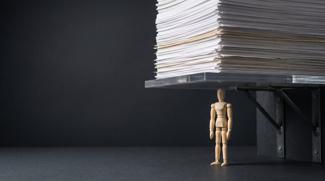 Small wooden mannequin feeling overwhelmed under heavy paper stack representing workplace stress, burnout and demanding office work load