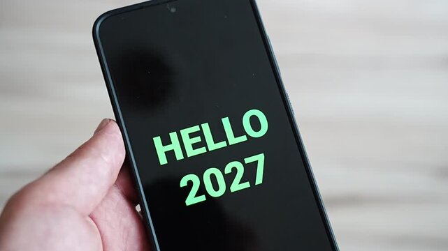 Close up of a tilted smartphone with Hello 2027 in large mint green letters, held by a man against a blurred light background. A bold, readable greeting for New Year Eve countdowns, upcoming year pro