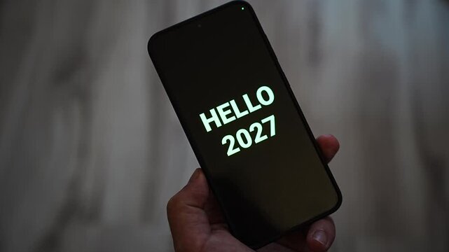 Hello 2027 greeting appears in glowing mint green letters on a black smartphone screen held in one hand. A message welcoming the new year, suited to New Year Eve wishes, fresh start and happy new yea