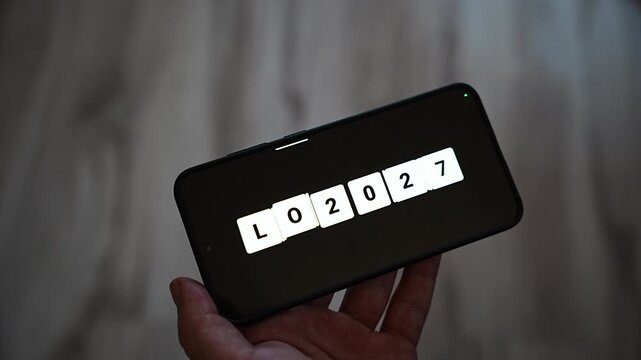 Loading 2027 spelled out tile by tile in white letter blocks on a smartphone held horizontally in one hand. A playful countdown to the coming year for New Year Eve, planning, anticipation and next ye