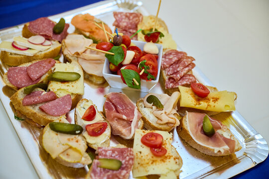 A catering tray filled with a variety of party snacks, including cold cut canapes, salami and ham bruschetta with gherkins, cherry tomatoes, cheese, and caprese skewers for a buffet event.
