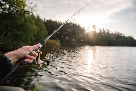 Person holding a modern spinning rod with a golden reel above a forest lake, angling from a boat during a bright sunset.