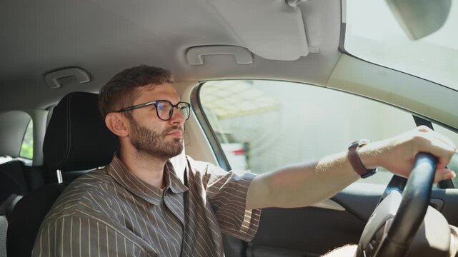 Young man driving car on sunny day, focused male driver with glasses holding steering wheel while commuting through green countryside on summer road trip