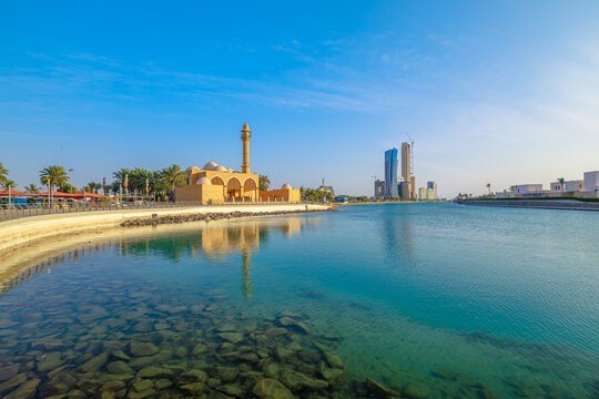 Serene waterfront with Masjid Abdul Qadir Faqih mosque reflecting on calm waters of Jeddah Corniche, with modern skyscrapers rising in distance under a clear blue sky at sunset in Jeddah, Saudi Arabia