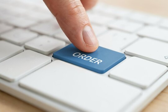 A finger presses a blue order button on a white computer keyboard during an online purchase.